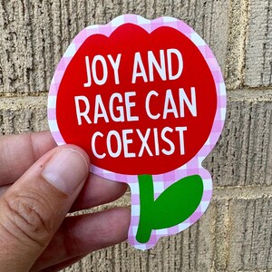 Peut inclure: Un autocollant en forme de fleur avec un centre rouge et une bordure rose et blanche &agrave; carreaux. L'autocollant porte les mots "JOY AND RAGE CAN COEXIST" en texte blanc. Une tige et une feuille vertes en bas.
