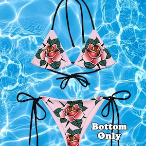 May include: A light pink bikini set featuring a traditional rose tattoo graphic with red petals, yellow centers, and dark green leaves. The top has triangle cups and black tie straps, while the bottom has side ties and the text Bottom Only.