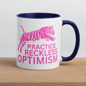 Può includere: Tazza in ceramica bianca con interno e manico blu navy. La tazza presenta una grafica di tigre rosa che salta e le parole "PRACTICE RECKLESS OPTIMISM" in rosa. La tazza è appoggiata su una superficie di legno.