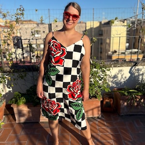 May include: A sleeveless knee-length dress featuring a black and white checkered pattern with large red rose graphics and green leaves. The dress has thin spaghetti straps and a V-neckline, paired with bright red framed sunglasses.