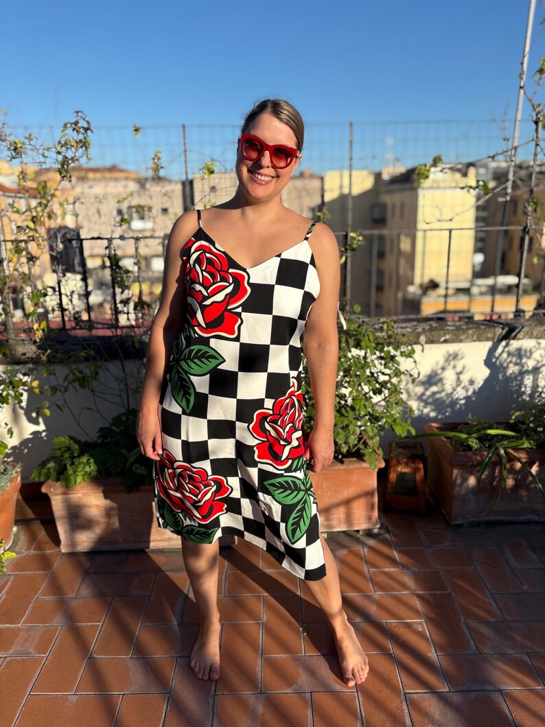 May include: A sleeveless knee-length dress featuring a black and white checkered pattern with large red rose graphics and green leaves. The dress has thin spaghetti straps and a V-neckline, paired with bright red framed sunglasses.