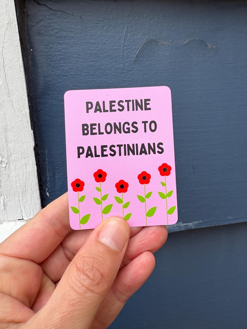 Może przedstawiać: R&oacute;żowy naklejka z tekstem "Palestine belongs to Palestinians" czarnym drukiem. Pięć czerwonych kwiat&oacute;w z zielonymi łodygami jest ułożonych u dołu naklejki.