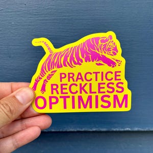 Può includere: Un adesivo giallo con un'illustrazione di tigre rosa e la scritta "PRACTICE RECKLESS OPTIMISM". L'adesivo è tenuto su uno sfondo blu. La tigre è raffigurata mentre salta.