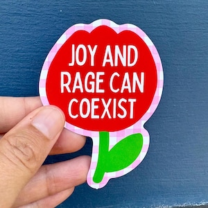Peut inclure: Un autocollant en forme de fleur avec un centre rouge et une bordure &agrave; carreaux roses et blancs. Le texte blanc "JOY AND RAGE CAN COEXIST" est inscrit sur l'autocollant. Une main tient l'autocollant.