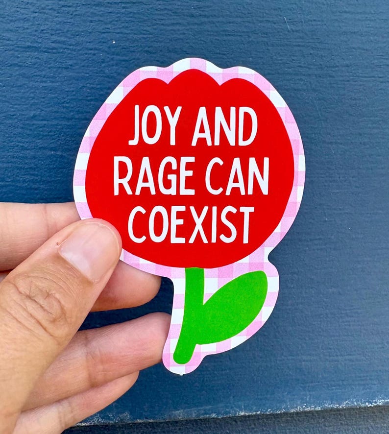 Peut inclure: Un autocollant en forme de fleur avec un centre rouge et une bordure &agrave; carreaux roses et blancs. Le texte blanc "JOY AND RAGE CAN COEXIST" est inscrit sur l'autocollant. Une main tient l'autocollant.