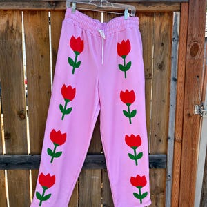 May include: Light pink sweatpants featuring a repeating vertical pattern of red tulips with green stems and leaves on each leg. The sweatpants have an elastic waistband with a white drawstring cord and are displayed on a clear plastic hanger.