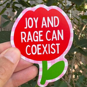 Peut inclure: Un autocollant rouge en forme de fleur avec le texte "JOY AND RAGE CAN COEXIST" en blanc. L'autocollant a une bordure rose et blanche &agrave; carreaux et des feuilles vertes. L'autocollant est tenu devant un fond de feuilles vertes.
