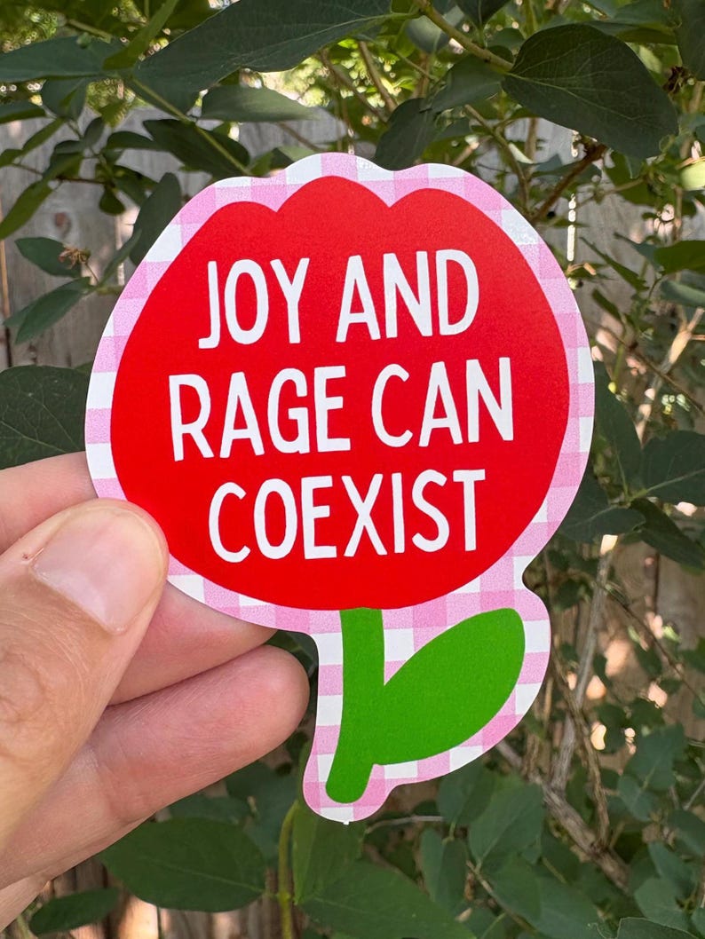 Peut inclure: Un autocollant rouge en forme de fleur avec le texte "JOY AND RAGE CAN COEXIST" en blanc. L'autocollant a une bordure rose et blanche &agrave; carreaux et des feuilles vertes. L'autocollant est tenu devant un fond de feuilles vertes.