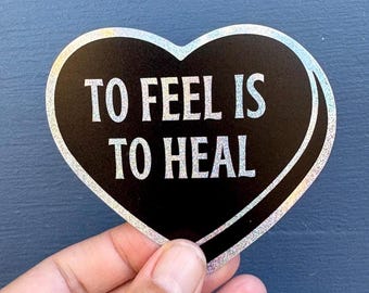 May include: A heart-shaped sticker with the text "TO FEEL IS TO HEAL" in white, outlined in silver glitter, against a black background. The sticker is held by a hand.