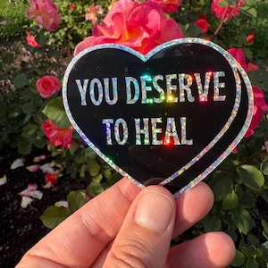 Könnte beinhalten: Ein schwarzer herzförmiger Aufkleber mit schimmerndem Glitzer. Der Aufkleber sagt "You Deserve to Heal" in weißer Schrift.