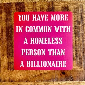 Może przedstawiać: Kwadratowy, jaskraworóżowy podkładka pod szklankę z białym tekstem: "YOU HAVE MORE IN COMMON WITH A HOMELESS PERSON THAN A BILLIONAIRE". Podkładka leży na drewnianej powierzchni.
