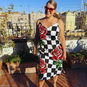May include: A sleeveless midi dress featuring a black and white checkered pattern with large red rose graphics and green leaves. The dress has thin spaghetti straps and a V-neckline. The garment is worn with red-framed sunglasses.