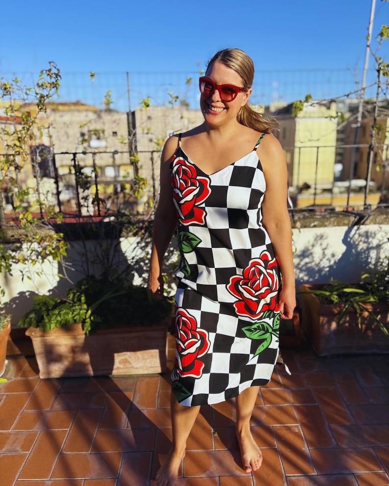 May include: A sleeveless midi dress featuring a black and white checkered pattern with large red rose graphics and green leaves. The dress has thin spaghetti straps and a V-neckline. The garment is worn with red-framed sunglasses.