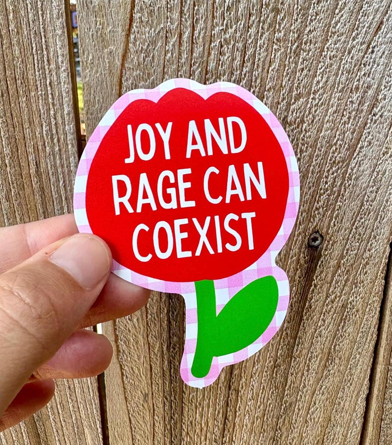 Peut inclure: Un autocollant en forme de fleur avec un centre rouge et le texte "JOY AND RAGE CAN COEXIST" en blanc. L'autocollant a une bordure vichy rose et blanche, une feuille verte, et est tenu sur un fond en bois.