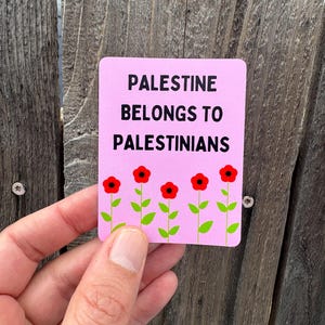 Może przedstawiać: R&oacute;żowy naklejka z tekstem "Palestine belongs to Palestinians" w czarnym kolorze. Pięć czerwonych kwiat&oacute;w z zielonymi łodygami jest ułożonych u dołu naklejki.