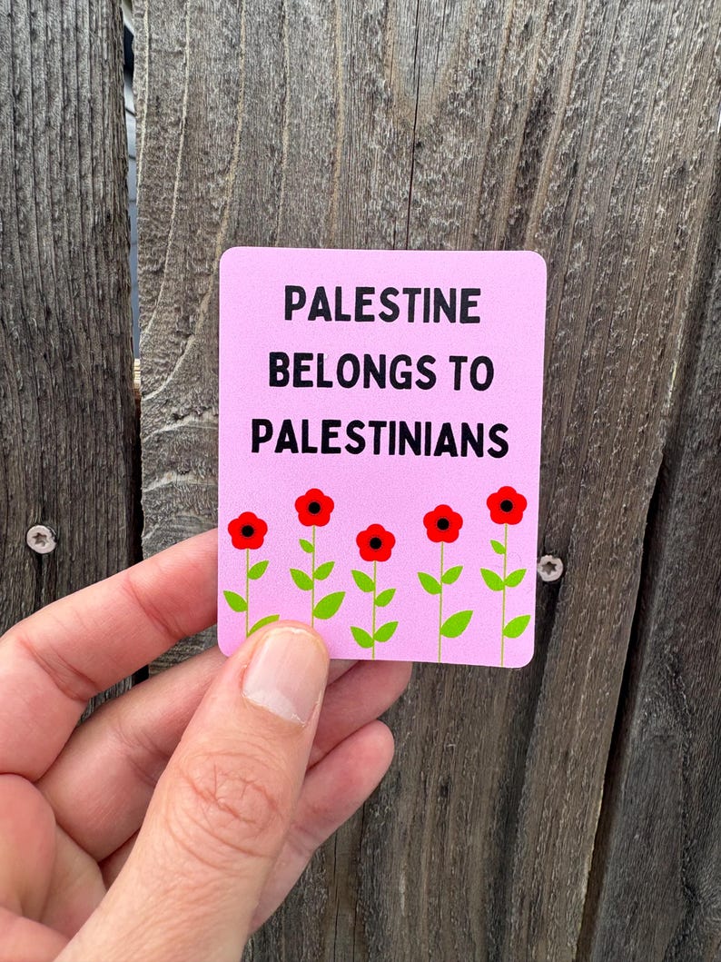 Może przedstawiać: R&oacute;żowy naklejka z tekstem "Palestine belongs to Palestinians" w czarnym kolorze. Pięć czerwonych kwiat&oacute;w z zielonymi łodygami jest ułożonych u dołu naklejki.