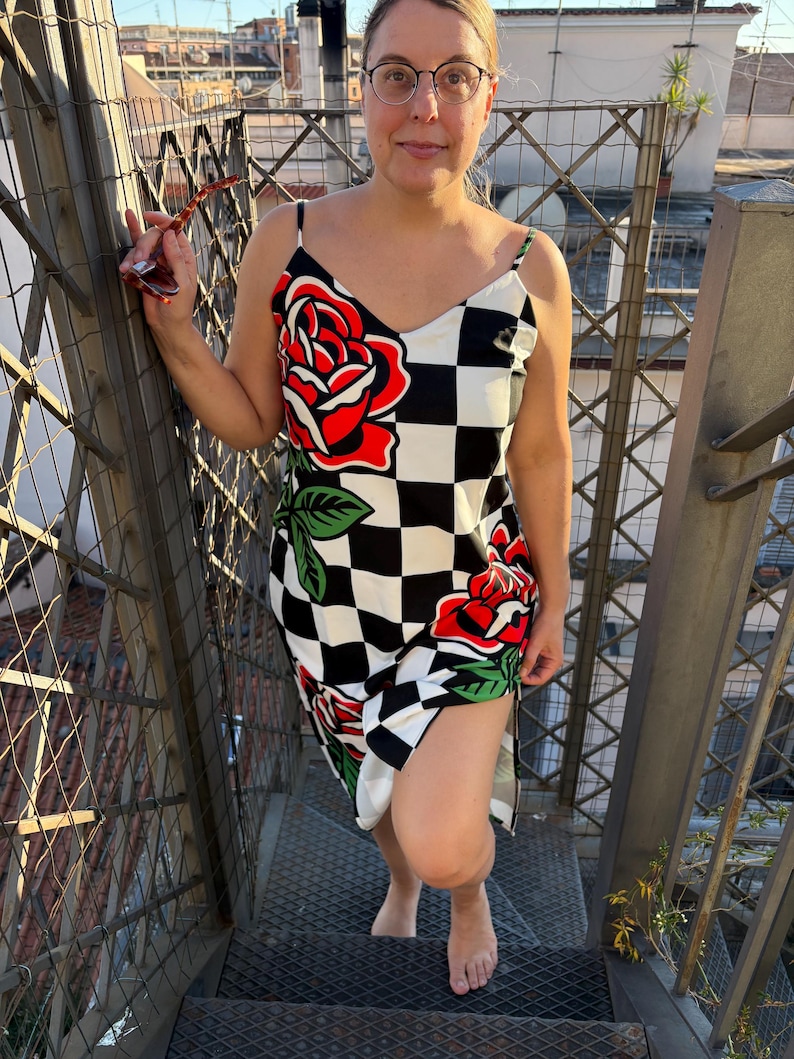 May include: A sleeveless slip dress featuring a black and white checkered pattern accented with large red rose graphics with green leaves. The dress has thin spaghetti straps and a V-neckline, falling to a mid-length hem with a side slit.