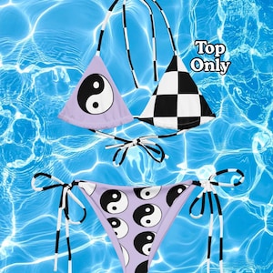 May include: A bikini set featuring a triangle top and tie-side bottoms. The top has one lavender cup with a black and white yin-yang symbol and one black and white checkered cup. The bottoms are lavender with a repeating yin-yang pattern. Text reads Top Only.