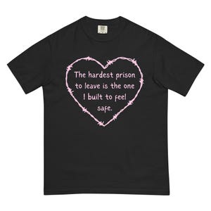 May include: A black t-shirt featuring a light pink heart outline made of barbed wire. Inside the heart, the text reads: The hardest prison to leave is the one I built to feel safe. The shirt has short sleeves and a crew neckline, presenting a graphic design.