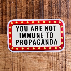 May include: A white sticker with a red border and yellow light bulbs around the edge. The sticker says "You are not immune to propaganda."