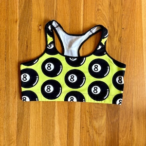 May include: A neon yellow racerback sports bra featuring a repeating pattern of black billiard eight balls with white numbers. The garment has a black trim along the neckline, armholes, and racerback straps, with a white interior lining visible.