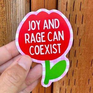 Peut inclure: Un autocollant en forme de fleur avec un centre rouge et une bordure vichy rose et blanche. Le texte blanc "JOY AND RAGE CAN COEXIST" est inscrit sur l'autocollant.
