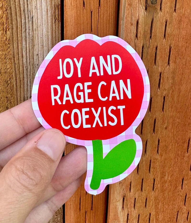 Peut inclure: Un autocollant en forme de fleur avec un centre rouge et une bordure vichy rose et blanche. Le texte blanc "JOY AND RAGE CAN COEXIST" est inscrit sur l'autocollant.