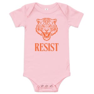 Può includere: Un body da bambino rosa chiaro con maniche corte. La parte anteriore presenta una grafica con la testa di una tigre arancione sopra la parola "RESIST" in lettere arancioni. Il body ha tre bottoni automatici sul fondo.