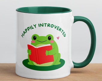 May include: White ceramic mug with a green rim and handle. The mug features a green frog reading a red book with the text "Happily Introverted".