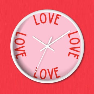 May include: A round wall clock with a white frame and a light pink face. The word LOVE is printed in red capital letters at the top, bottom, left, and right positions on the clock face. The clock features white hour, minute, and second hands.