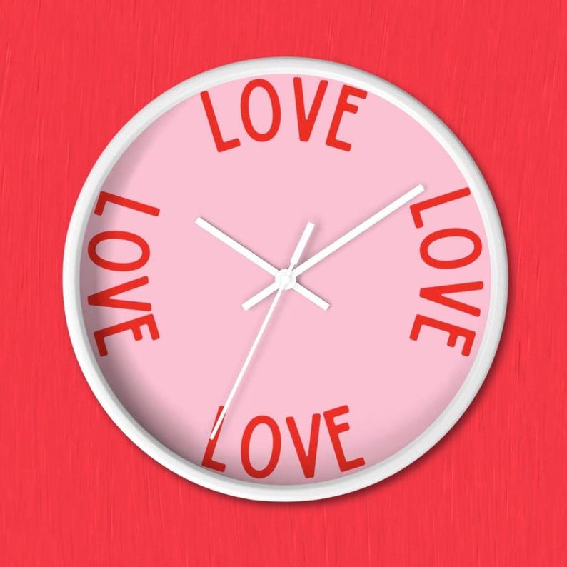 May include: A round wall clock with a white frame and a light pink face. The word LOVE is printed in red capital letters at the top, bottom, left, and right positions on the clock face. The clock features white hour, minute, and second hands.
