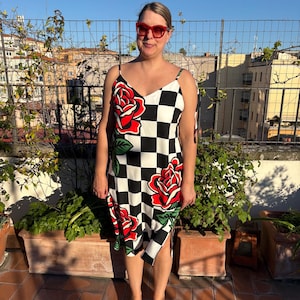 May include: A sleeveless midi dress featuring a black and white checkered pattern with large red rose graphics and green leaves. The dress has thin spaghetti straps, a V-neckline, and a side slit at the hem. Red-framed sunglasses are also worn.