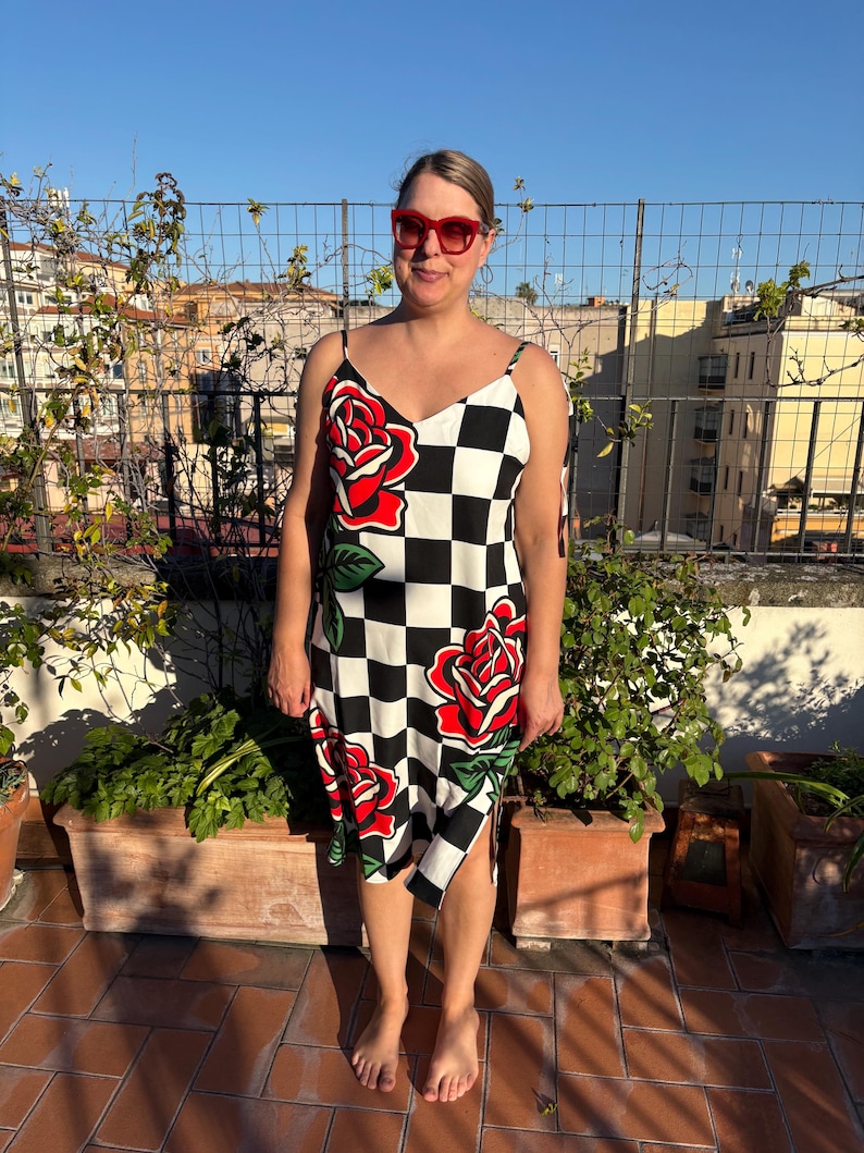 May include: A sleeveless midi dress featuring a black and white checkered pattern with large red rose graphics and green leaves. The dress has thin spaghetti straps, a V-neckline, and a side slit at the hem. Red-framed sunglasses are also worn.