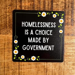May include: Black sign with white text and a daisy border. The text reads "Homelessness is a choice made by government."