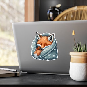 Peut inclure: Un adorable renard orange enroulé dans une couverture bleue avec des flocons de neige blancs. Le renard est blotti dans la couverture, les yeux fermés.