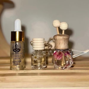 May include: Three car air fresheners. One is a clear glass bottle with a wooden top and a white string. The second is a clear glass bottle with a wooden top and dried flowers inside. The third is a small glass bottle with a gold cap and a label that reads "Earthbound Coconut Vanilla Essential Oil Refill".