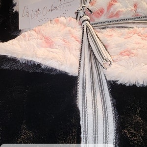 May include: A 60" x 90" black and white faux fur throw blanket with a pink and white flamingo print. The blanket is tied with a white and black striped ribbon. The text "Lil' Little Dinker Designs" is on a white tag. The text "60" x 90" Wild Lynx - Flamingo Sable - Black $200" is on a white tag.