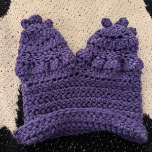 May include: A handmade, purple crochet hat with a unique, textured design. The hat features a close-fitting style with decorative elements at the top. The crochet work is visible, showcasing the yarn's texture and color.