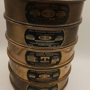 May include: A stack of five vintage brass sieves with black labels. Each sieve has a different mesh size, indicated by numbers and measurements in inches and millimeters. The sieves are from The W. S. Tyler Company.