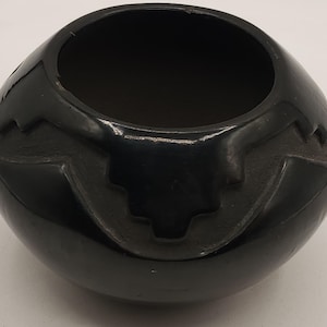 May include: A glossy black ceramic bowl with a rounded shape and a wide opening. The bowl features carved geometric designs on the sides. The interior is dark, contrasting with the smooth, reflective exterior.
