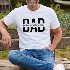 May include: White t-shirt with the word "DAD" in large, bold, black letters. Below the word "DAD" are the names "TARA", "EMMA", and "TOM" separated by stars. The person is wearing blue jeans and white sneakers.