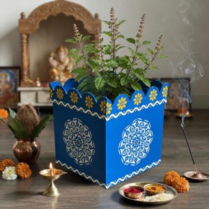 May include: A square blue planter with a scalloped top edge featuring yellow floral patterns and white mandala designs. The planter holds a green Tulsi plant. It is surrounded by a brass oil lamp, a copper pot with a coconut, and incense.