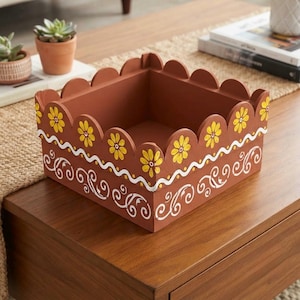 May include: A square terracotta-colored wooden box with a scalloped top edge. The exterior features hand-painted yellow flowers along the rim, a white wavy line, and white swirling scroll patterns on the sides of the container.
