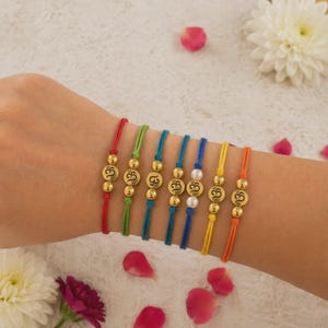 May include: Seven thin cord bracelets in red, green, teal, blue, yellow, and orange. Each bracelet features a central gold-toned circular charm embossed with an Om symbol, flanked by two small gold-toned beads. One blue bracelet includes two white pearls.