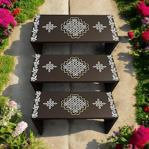 May include: Three dark brown wooden step stools with decorative white and gold geometric patterns. The stools are arranged in a descending staircase formation, with floral accents in the background.
