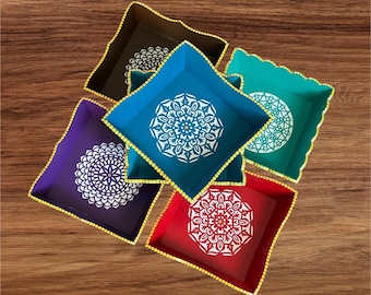 May include: Five square trays in dark brown, blue, teal, purple, and red, each featuring a white mandala pattern in the center and a gold beaded border along the edges. The trays are arranged in a cluster, showcasing their distinct colors.