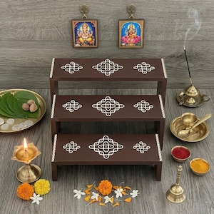 May include: A tiered, dark brown wooden display shelf with white geometric designs. The shelf is decorated with religious items, including small framed pictures, brass bowls, a bell, and incense. A lit oil lamp and flowers are also present.