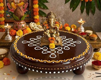 May include: A brown, round, wooden pedestal table with a decorative white and yellow border and a white geometric design. A gold statue of Ganesha sits on the table, surrounded by flowers and candles. The background includes a framed image of Ganesha and floral garlands.