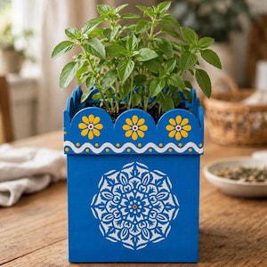 May include: A square blue wooden planter featuring a white mandala pattern on the front, a scalloped top edge decorated with yellow flowers, and a white wavy line detail. A green basil plant grows from the top of the container.