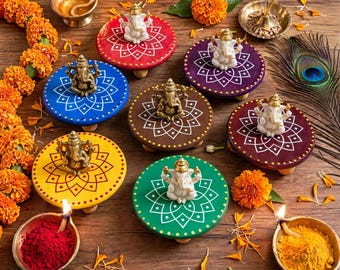 May include: Seven small Ganesha figurines placed on colorful circular wooden stands with white mandala patterns. The set includes red, blue, yellow, green, brown, purple, and maroon bases, surrounded by marigold flowers and bowls of colored powder.